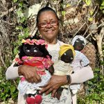 Guillot Ramos smiles, standing outside holding three smiling Black dolls wearing dresses and head scarves