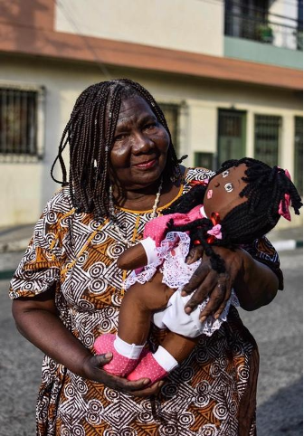 Grueso lleva un vestido con estampado geométrico y acuna un paño, muñeca negra de peluche con un traje rosa