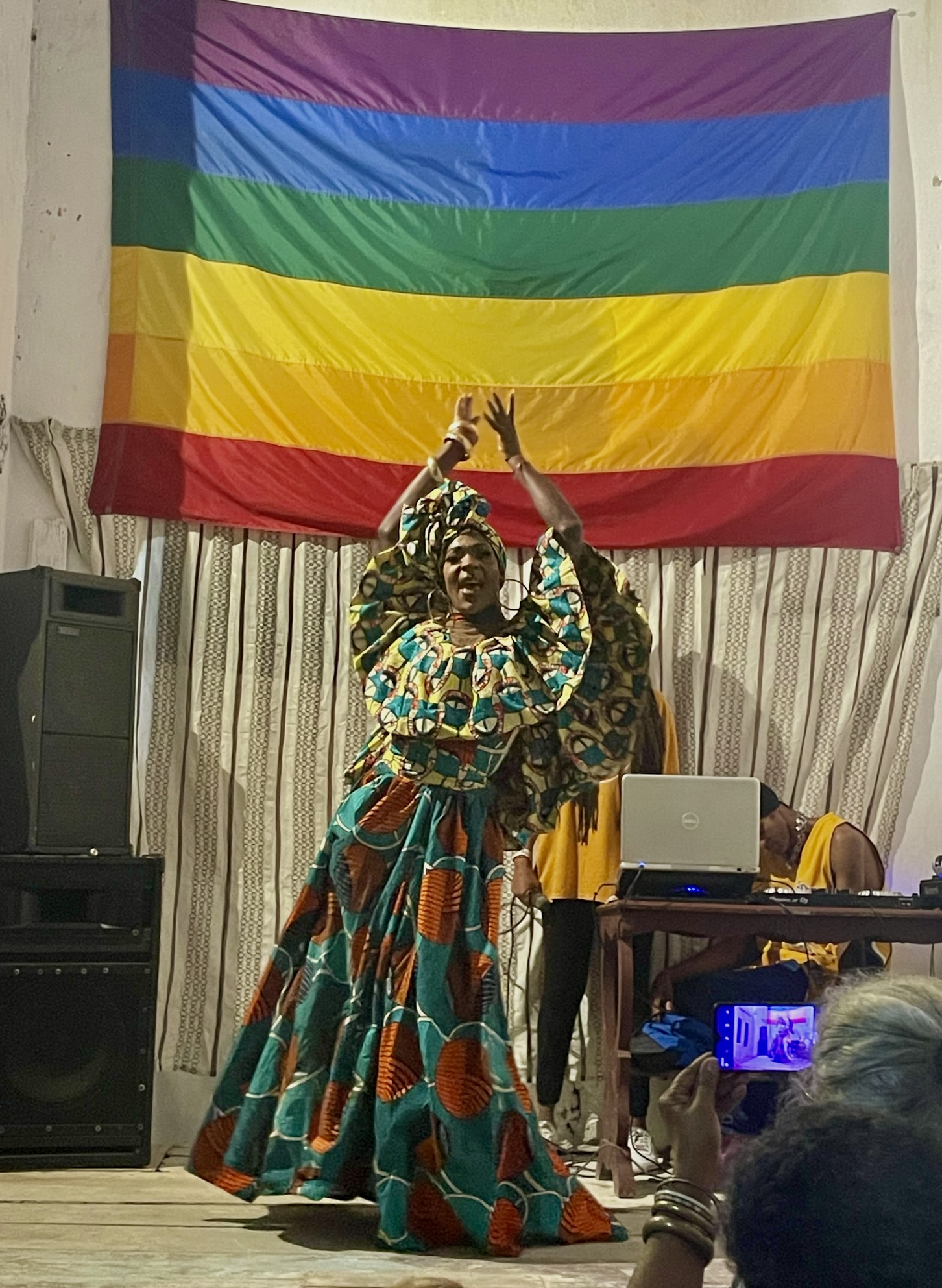 Una persona con un vestido de colores y un pañuelo en la cabeza baila con los brazos por encima de la cabeza frente a una gran bandera LGBTQ.